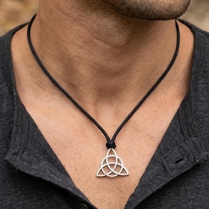 May include: A silver-toned triquetra pendant suspended from a thin black cord. The pendant features an interlaced three-pointed knot design with a polished metallic finish. The black cord is secured around the neck, holding the pendant centered.