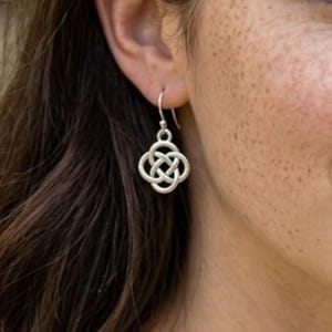 May include: A silver-toned dangle earring featuring a four-lobed Celtic knot design. The charm is suspended from a simple curved ear wire hook, showcasing a metallic finish with intricate, interconnected loops forming a symmetrical pattern.