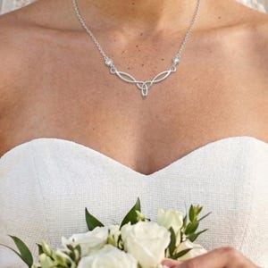 May include: A silver-toned necklace featuring a central trinity knot design with two delicate, curved metal branches extending outward, each accented by a single small, clear round gemstone set into the chain on either side of the pendant.