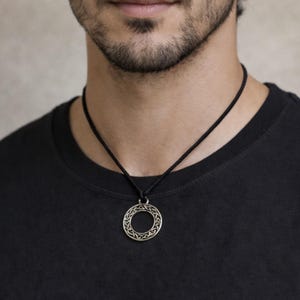 May include: A silver-colored pendant necklace with a circular design. The pendant hangs from a black cord. The necklace is worn against a black shirt.