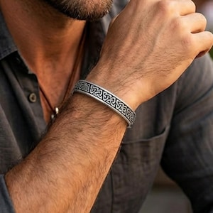 May include: A silver-toned metal cuff bracelet featuring a continuous engraved pattern of interlocking triangular knots. The band has a polished finish with darkened recessed areas that highlight the intricate geometric design across the surface.