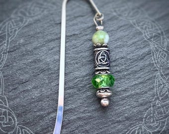 Op de afbeelding: Een zilverkleurige metalen bladwijzer met een decoratieve hanger. De hanger bestaat uit een lichtgroene ronde kraal, een zilverkleurige cilindrische kraal met een triquetra-knoopmotief en een gefacetteerde felgroene glaskraal.