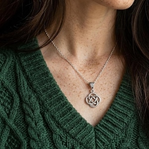 Op de afbeelding: Een zilverkleurige metalen ketting met een hanger in de vorm van een ronde Keltische knoop. De hanger hangt aan een eenvoudige zilverkleurige ketting. Het ingewikkelde, verweven patroon vormt een doorlopende lus aan een oogje.