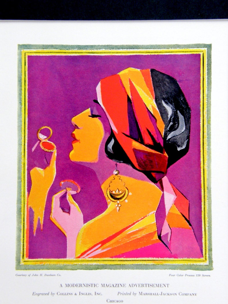 May include: A woman with dark hair is wearing a red, yellow, and black turban. She is holding a compact mirror in her right hand and applying makeup with her left hand. The background is a solid purple color.