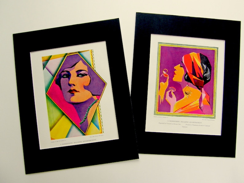 May include: Two framed vintage art prints featuring colorful illustrations of women in geometric designs. The first print features a woman with purple hair and a pink and yellow background. The second print features a woman with a red and orange turban and a purple background. Both prints have a black mat.
