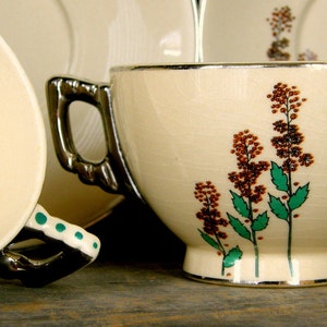 Puede incluir: Un juego de taza y platillo vintage con un diseño floral. La taza tiene un borde plateado y un asa con un acento verde. El platillo tiene un fondo blanco con un patrón floral.