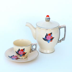 Puede incluir: Un juego de té vintage con una tetera de color crema y una taza y un platillo a juego. La tetera tiene un pomo rojo en la tapa y un diseño floral en tonos rojos, azules y negros. La taza y el platillo tienen un diseño floral similar.