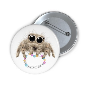 Op de afbeelding: Een witte button met een cartoon spin. De spin heeft grote ogen en een pluizig lichaam en houdt een armband met het woord "BESTIES" vast. Een tweede, zilveren button is zichtbaar op de achtergrond.