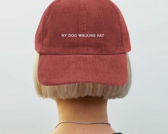 May include: A brick-red corduroy baseball cap with the text "MY DOG WALKING HAT" in white. The hat has a curved brim and a rounded top. The image shows the back of a person wearing the hat.