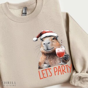 May include: Beige sweatshirt featuring a capybara wearing a Santa hat and holding a cocktail. The capybara is smiling, and the words "LET'S PARTY" are printed in red. The sweatshirt has a crew neck and long sleeves.