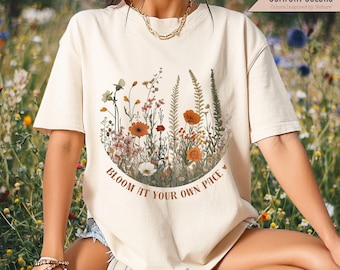 Puede incluir: Una camiseta beige con un diseño floral y el texto "Bloom at your own pace" en letras marrones. La camiseta tiene un corte holgado y mangas cortas.