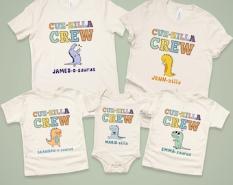 May include: Five off-white t-shirts and a onesie with the text "CUZ-ZILLA CREW" in colorful letters. Each shirt features a different dinosaur illustration and a name: James-o-saurus, Jenn-zilla, Brandon-o-saurus, Marie-zilla, and Emma-saurus.