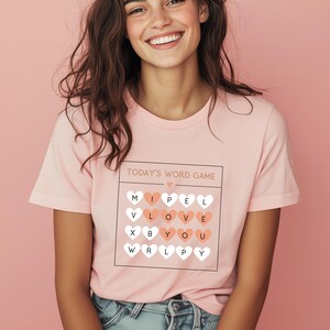 May include: A pink t-shirt with a white graphic that says "TODAY'S WORD GAME" and features a grid of hearts with letters inside. The letters spell out "I LOVE YOU".