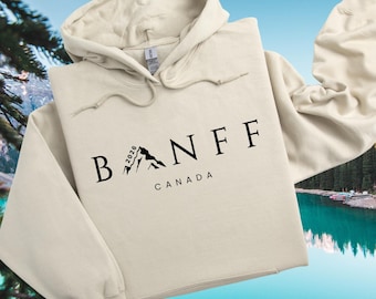 May include: A cream-colored hoodie with the word "BANFF" in black capital letters, with a mountain graphic and the word "CANADA" below. The year "2026" is printed on the mountain graphic. The hoodie has a drawstring hood.