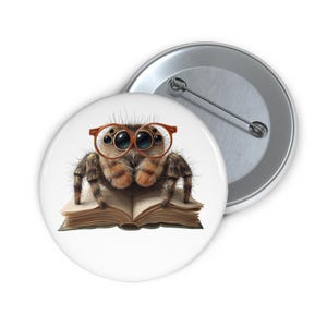 Op de afbeelding: Een witte button met een cartoon spin met een oranje bril die een boek leest. De spin is bruin en bruingeel met gedetailleerde vacht. Een tweede, zilveren button is zichtbaar op de achtergrond.