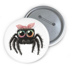 Op de afbeelding: Een witte button met een cartoon spin. De zwarte, harige spin heeft grote, kleurrijke ogen en een roze strik. Een tweede button met een zilveren speld is ook zichtbaar.