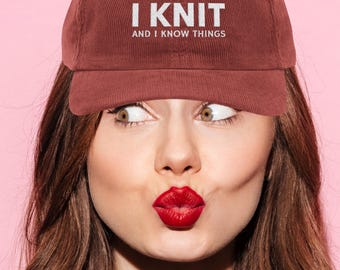 Może przedstawiać: Bordowa sztruksowa czapka z daszkiem z białym napisem "I KNIT AND I KNOW THINGS". Czapkę nosi osoba z czerwoną szminką i brązowymi włosami na różowym tle.