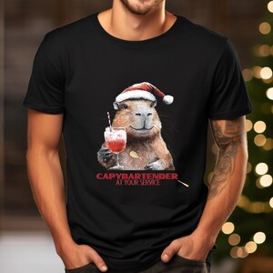 May include: Black t-shirt featuring a capybara wearing a Santa hat and holding a cocktail. The capybara has a spoon and the text "CAPYBARTENDER AT YOUR SERVICE" in red. The shirt is a crew neck.