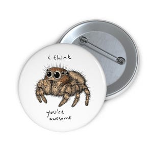 Op de afbeelding: Een witte button met een cartoon spin en de tekst "i think you're awesome". De spin is bruin en beige met gedetailleerde schaduwen. Een tweede, zilveren button is zichtbaar op de achtergrond.