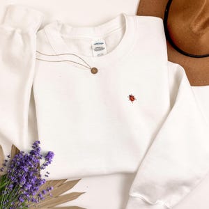 May include: White sweatshirt with a small embroidered ladybug design. The sweatshirt has a crew neck and long sleeves.