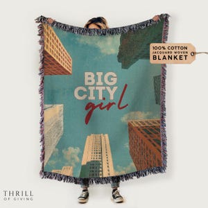 May include: A woven throw blanket with a blue background and a cityscape design. The text "BIG CITY girl" is printed in red and white on the blanket. The blanket is made of 100% cotton.