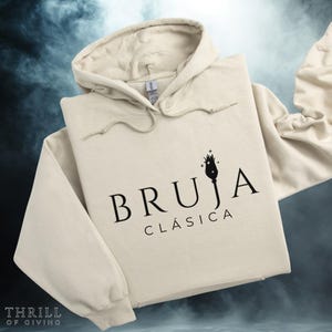 Puede incluir: Una sudadera con capucha beige con la inscripción "BRUJA CLÁSICA" en negro. El diseño incluye una ilustración estilizada de un sombrero de bruja con estrellas. La sudadera tiene una capucha con cordón y un bolsillo delantero.