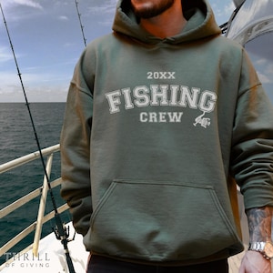Puede incluir: Sudadera con capucha verde oliva con el texto "FISHING CREW" en letras blancas. La sudadera tiene un bolsillo delantero y un gráfico de caña de pescar. El año "20XX" está encima del texto. La imagen está tomada en un barco.