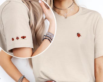 Puede incluir: Camiseta beige con una pequeña mariquita roja bordada en el pecho. La manga izquierda presenta tres mariquitas bordadas. La camiseta es de color neutro y tiene un diseño clásico de cuello redondo.