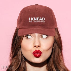 Puede incluir: Gorra de béisbol de pana granate con el texto "I KNEAD AND I KNOW THINGS" en blanco. La gorra la lleva una persona con pintalabios rojo y pelo castaño, sobre un fondo rosa. La esquina inferior izquierda tiene el texto "THRILL OF GIVING".