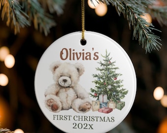 May include: A white ceramic Christmas ornament with a teddy bear and Christmas tree design. The text "Olivia's First Christmas 202X" is printed on the ornament. The ornament is hanging from a gold string.