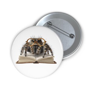 Op de afbeelding: Een witte knoop met een gedetailleerde illustratie van een spin met een ronde, goudkleurige bril, die op een open boek zit. De spin is bruin en grijs met een harige textuur. Een tweede knoop is zichtbaar op de achtergrond.