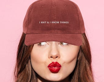 Może przedstawiać: Bordowa sztruksowa czapka bejsbolówka z białym napisem "I KNIT & I KNOW THINGS". Czapka jest noszona przez osobę z czerwoną szminką i brązowymi włosami na różowym tle.