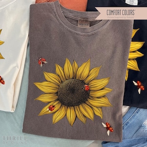 May include: A brown t-shirt with a sunflower graphic and red ladybugs. The shirt has the text "COMFORT COLORS" and "Colors Inspired by Nature" printed on it.