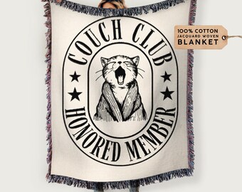 May include: A white woven throw blanket with a black and white illustration of a cat wearing a robe. The illustration is inside a circle with the text "Couch Club Honored Member" around the outside. The text "100% Cotton Jacquard Woven Blanket" is in a brown rectangle in the top right corner.