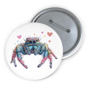 Op de afbeelding: Een witte button met een kleurrijke illustratie van een springspin. De spin heeft een blauw lichaam, roze en gele poten en meerdere ogen. Kleine hartjes in roze, geel en oranje zijn ook afgebeeld.