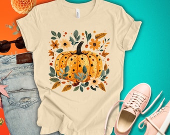 May include: A beige t-shirt with a graphic of a yellow pumpkin with brown spots surrounded by orange, brown, and green leaves and flowers.
