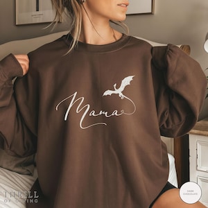 May include: A dark chocolate brown sweatshirt featuring the word "Mama" in white script, with a white dragon illustration. The sweatshirt has a crew neck and long sleeves. The text "Thrill of Giving" is at the bottom left.