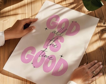 May include: A cream-colored poster with the words "GOD is all the time GOOD" in pink and black. The poster is held by two hands on a wooden surface. The text is in a playful, retro font.