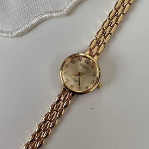 May include: A gold-toned wristwatch with a round face and a linked band. The watch face displays the numbers 1 through 12. The watch is lying on a white surface next to a cup of coffee.