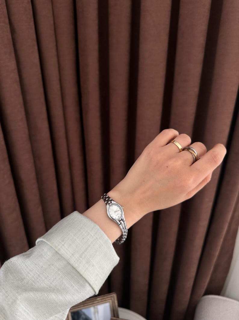 Op de afbeelding: Een zilveren polshorloge met een witte wijzerplaat en een schakelband wordt getoond. Het horloge wordt om een pols gedragen, met een lichtgroene mouw zichtbaar. De achtergrond bestaat uit bruine gordijnen.
