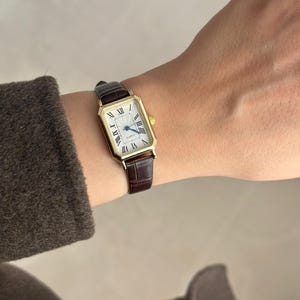 May include: A gold rectangular wristwatch with a white face and black Roman numerals. The watch has a brown leather band. The watch is on a wrist.