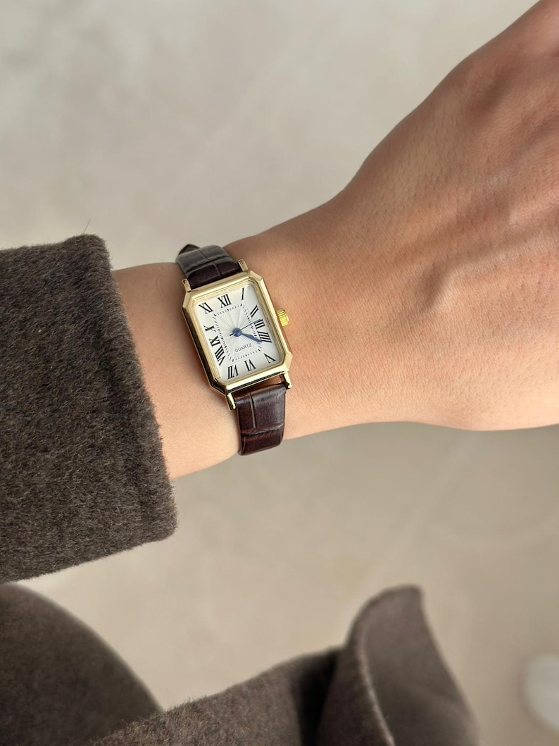 May include: A gold rectangular wristwatch with a white face and black Roman numerals. The watch has a brown leather band. The watch is on a wrist.