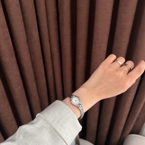 Op de afbeelding: Een zilveren polshorloge met een witte wijzerplaat en een schakelband wordt om een pols gedragen. Het horloge staat centraal, met een lichtgroene mouw en gouden ringen op de vingers op de achtergrond. Een klassiek, elegant accessoire.