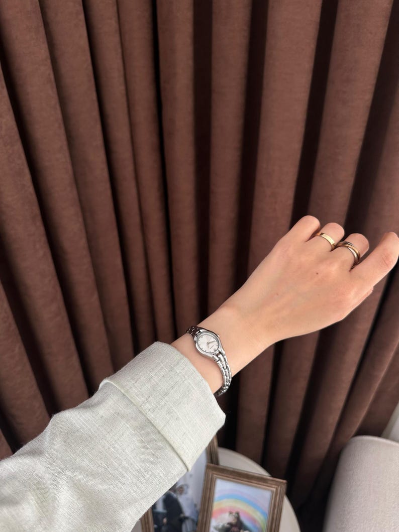 Op de afbeelding: Een zilveren polshorloge met een witte wijzerplaat en een schakelband wordt om een pols gedragen. Het horloge staat centraal, met een lichtgroene mouw en gouden ringen op de vingers op de achtergrond. Een klassiek, elegant accessoire.