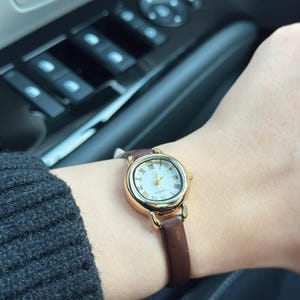 May include: A gold-toned quartz watch with a white face and Roman numeral hour markers. The watch has a brown leather band. The watch is on a wrist.
