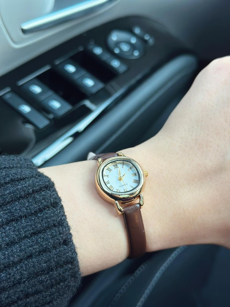 May include: A gold-toned quartz watch with a white face and Roman numeral hour markers. The watch has a brown leather band. The watch is on a wrist.