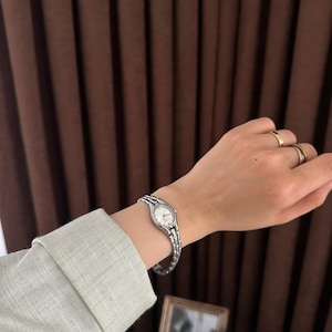Op de afbeelding: Een zilverkleurig polshorloge met een witte wijzerplaat wordt om een pols gedragen. Het horloge heeft een schakelband. De persoon draagt een lichtgroen shirt met lange mouwen. Twee ingelijste foto's staan op de achtergrond.