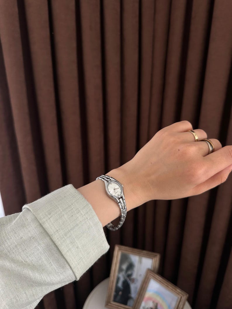 Op de afbeelding: Een zilverkleurig polshorloge met een witte wijzerplaat wordt om een pols gedragen. Het horloge heeft een schakelband. De persoon draagt een lichtgroen shirt met lange mouwen. Twee ingelijste foto's staan op de achtergrond.