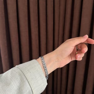 Op de afbeelding: Een zilverkleurige armband met een rechthoekig schakelontwerp. De armband wordt om een pols gedragen, met een lichtgroen shirt met lange mouwen zichtbaar. De achtergrond heeft bruine gordijnen.