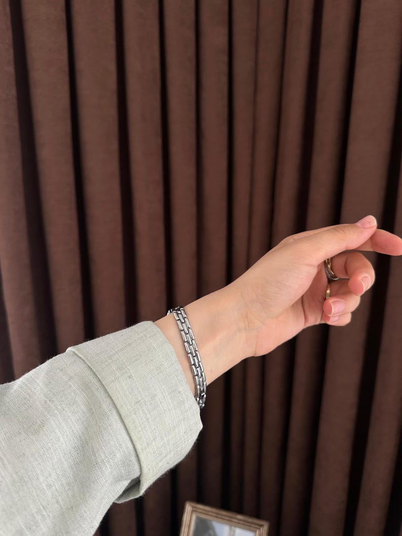 Op de afbeelding: Een zilverkleurige armband met een rechthoekig schakelontwerp. De armband wordt om een pols gedragen, met een lichtgroen shirt met lange mouwen zichtbaar. De achtergrond heeft bruine gordijnen.